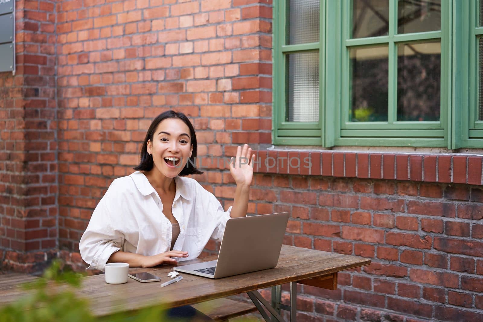 Friendly asian woman sitting with laptop, waving at you, saying hello, hi gesture, greeting you while working with computer.