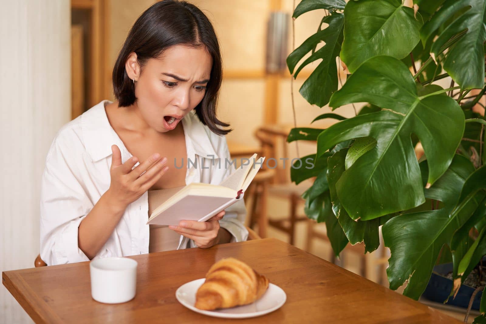 Portrait of asian woman looking shocked at book pages, reading something interesting, concentrating, sitting in cafe with coffee and croissant.