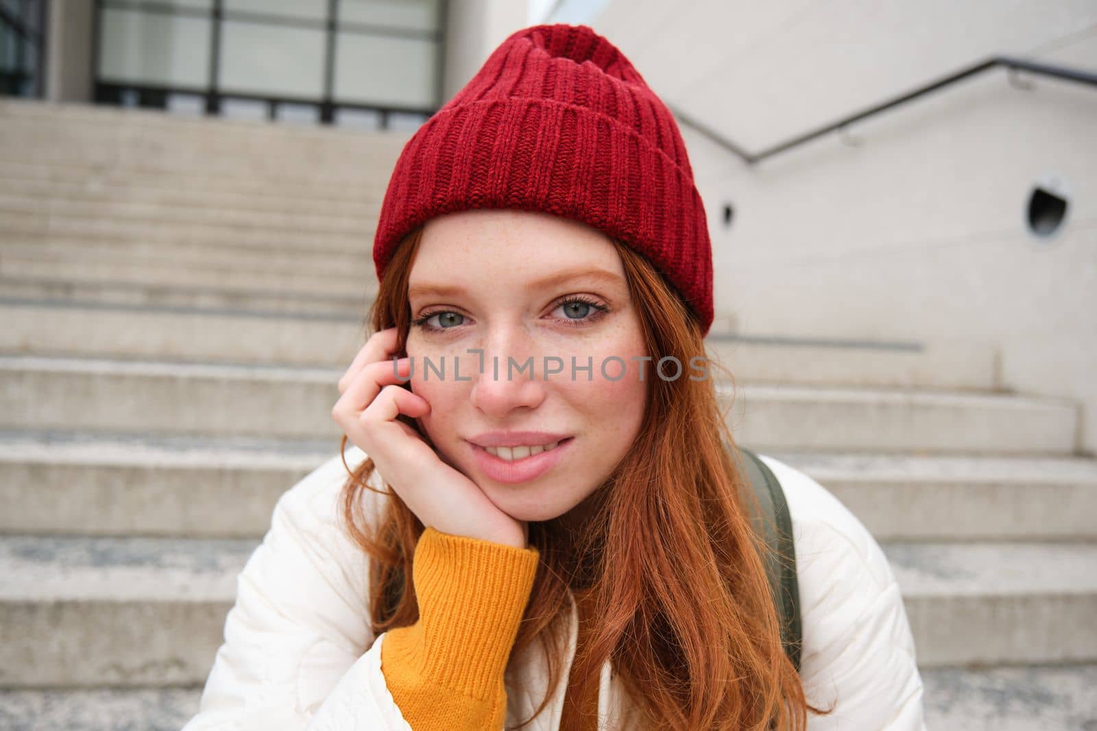 Close up portrait of beautiful redhead girl in red hat, urban woman with freckles and ginger hair, sits on stairs on street, smiles and looks gorgeous by Benzoix