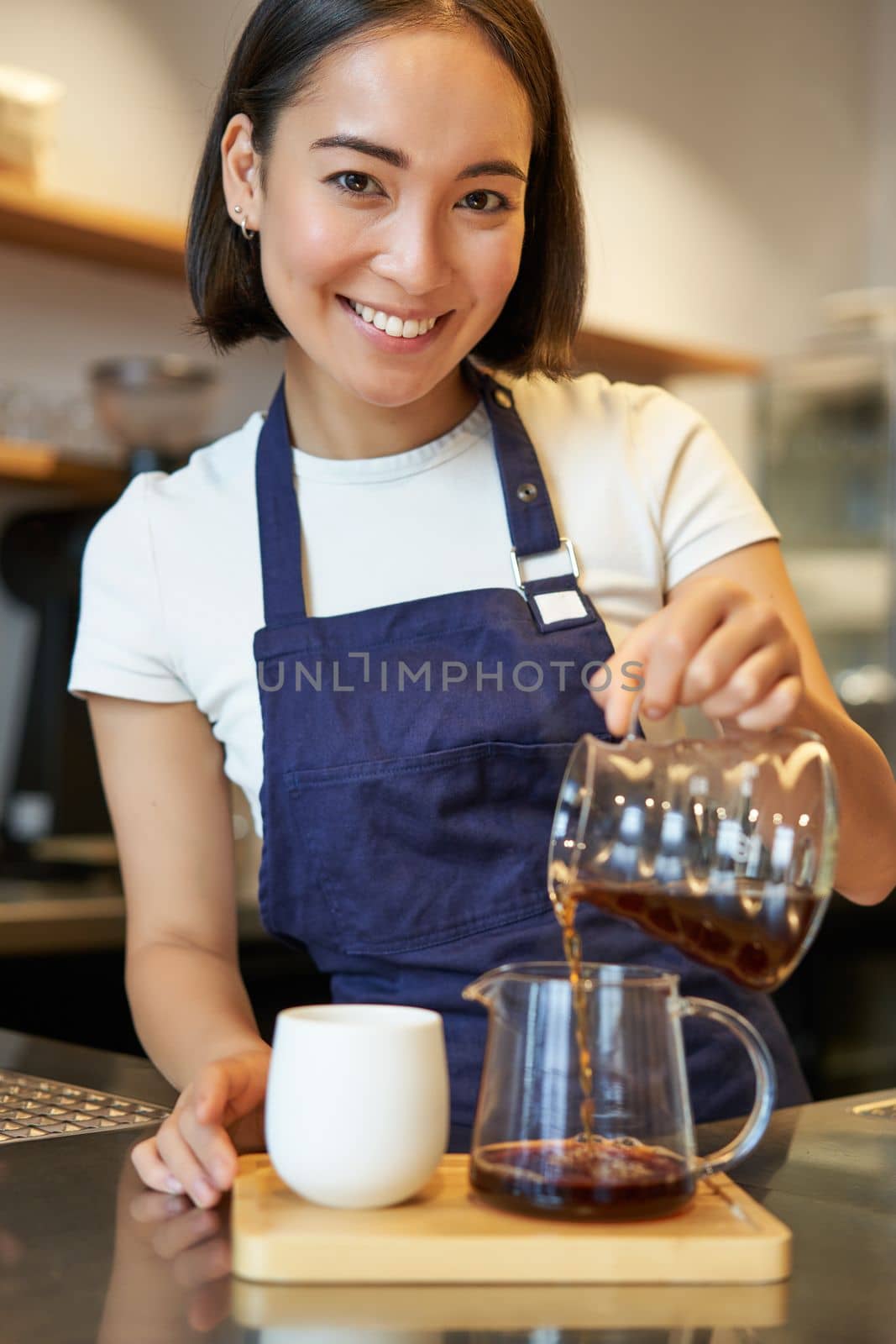 Vertical shot of smiling cute barista girl, pouring filter coffee, making order for cafe client, wearing blue apron by Benzoix