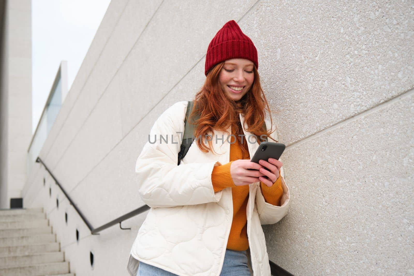 Redhead girl, young woman tourist with backpack, holds smartphone, looks for route on mobile application, searches for hotel on phone map, smiles happily by Benzoix