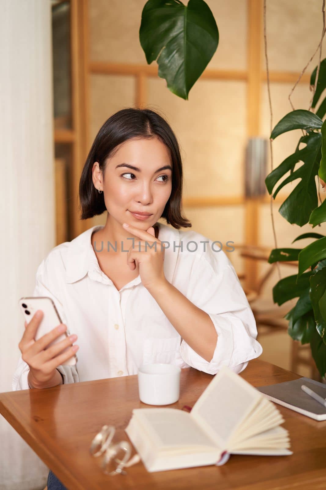 Beautiful young woman, 25 years old, thinking, holding smartphone and looking thoughtful, sitting in cafe, deciding smth by Benzoix