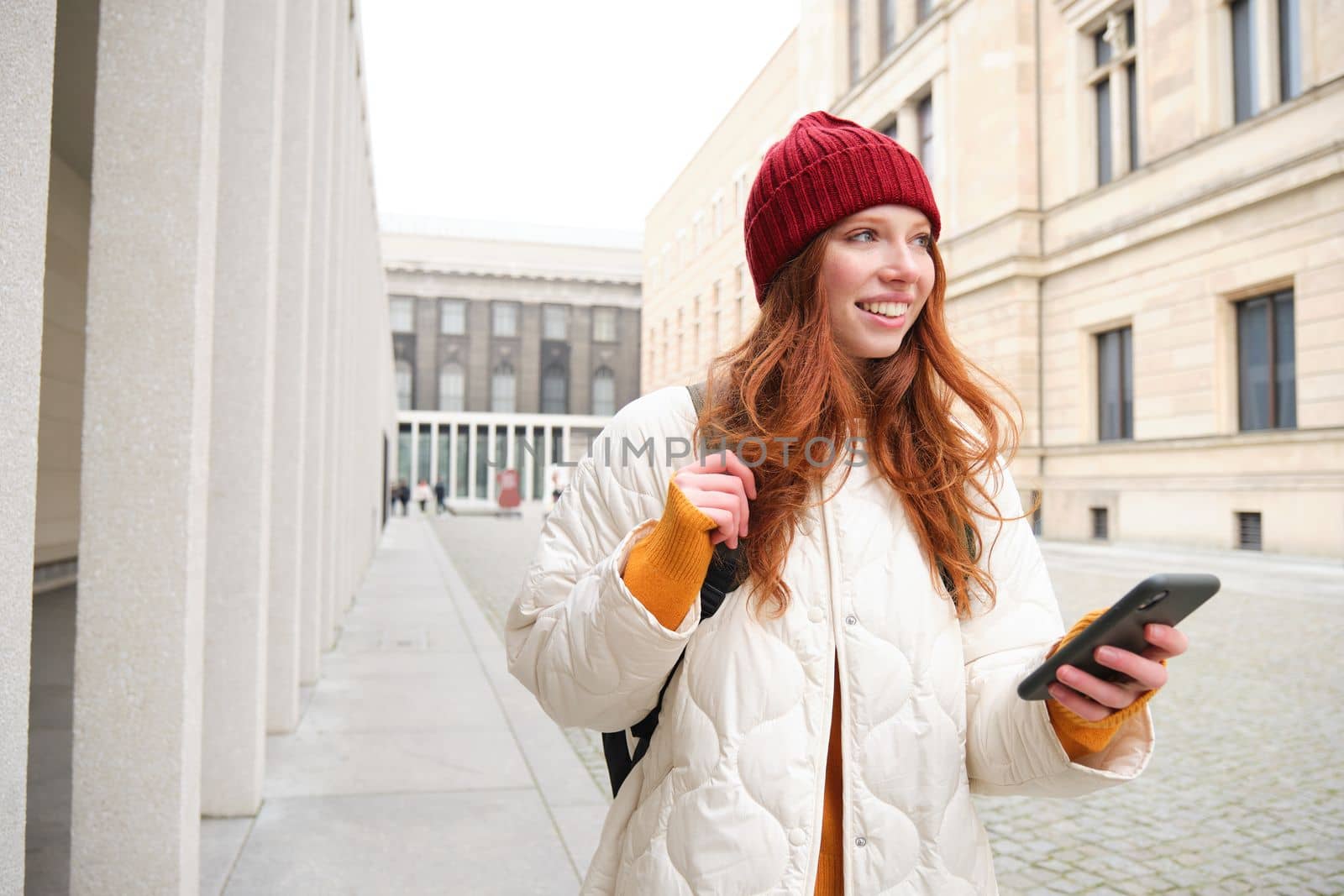Beautiful smiling girl, tourist with backpack, holding smartphone, using map on mobile phone application, looking for sightseeing in internet app, standing outdoors.