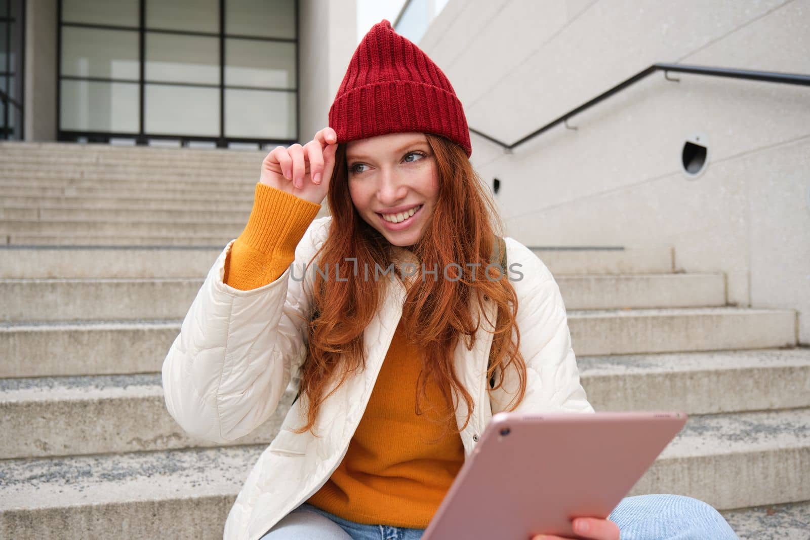 Beautiful redhead female model, ginger girl with digital tablet, sits on stairs outdoors, reads on her gadget, uses internet application and wifi on street by Benzoix