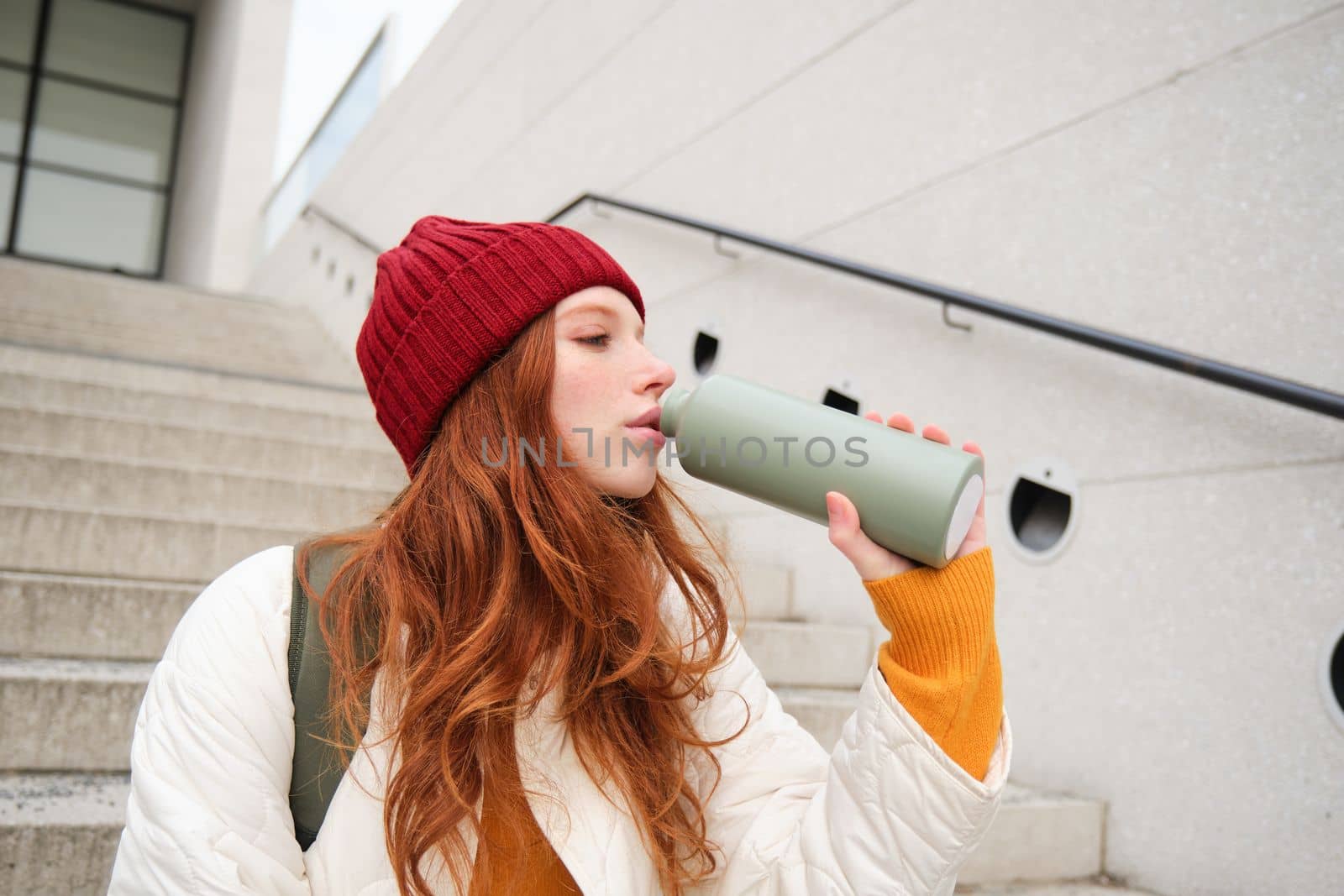 Smiling traveler, redhead girl tourist sits on stairs with flask, drinks hot coffee from thermos while travelling and sightseeing around foreign city, sits on stairs and rests by Benzoix