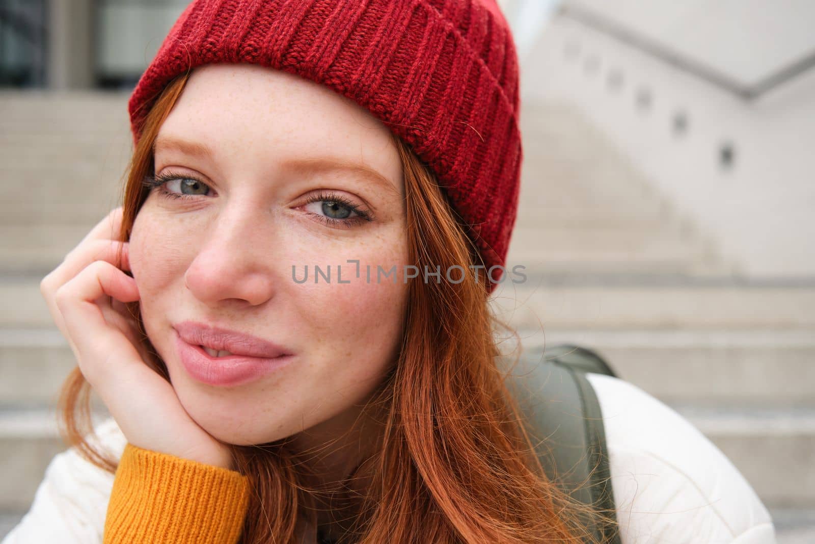 Close up portrait of beautiful redhead girl in red hat, urban woman with freckles and ginger hair, sits on stairs on street, smiles and looks gorgeous by Benzoix
