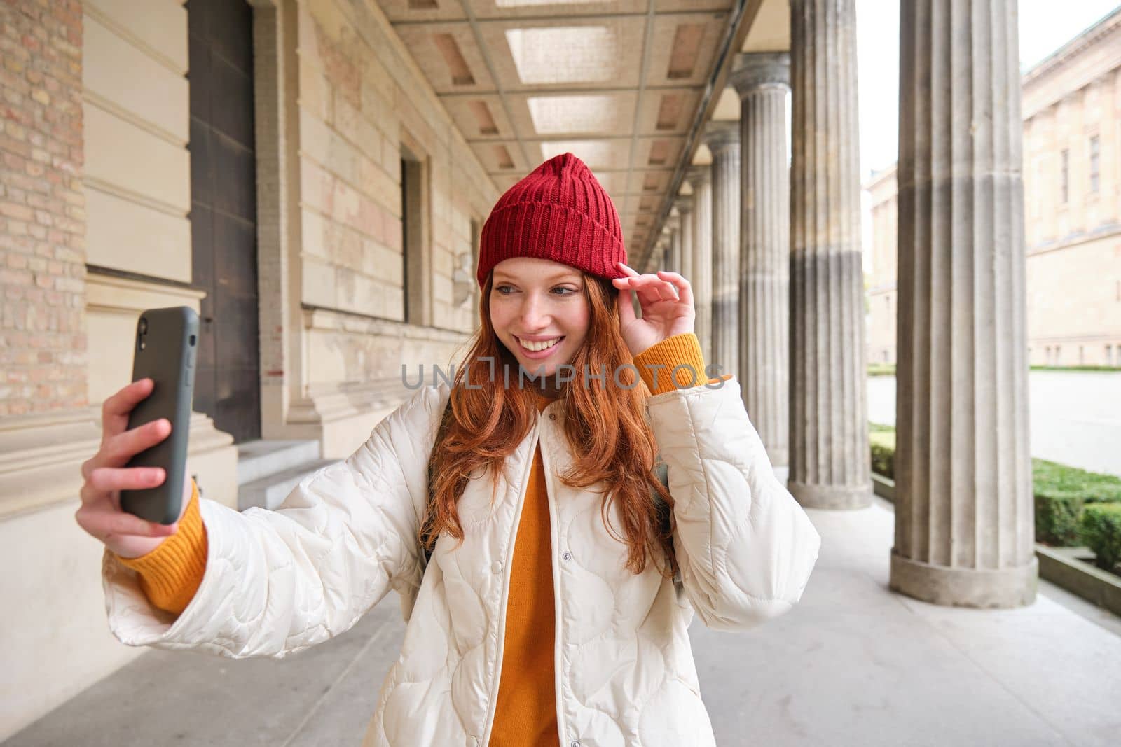 Stylish redhead girl tourist, takes selfie in front of tourism attraction, makes photo with smartphone, looks at mobile camera and poses.
