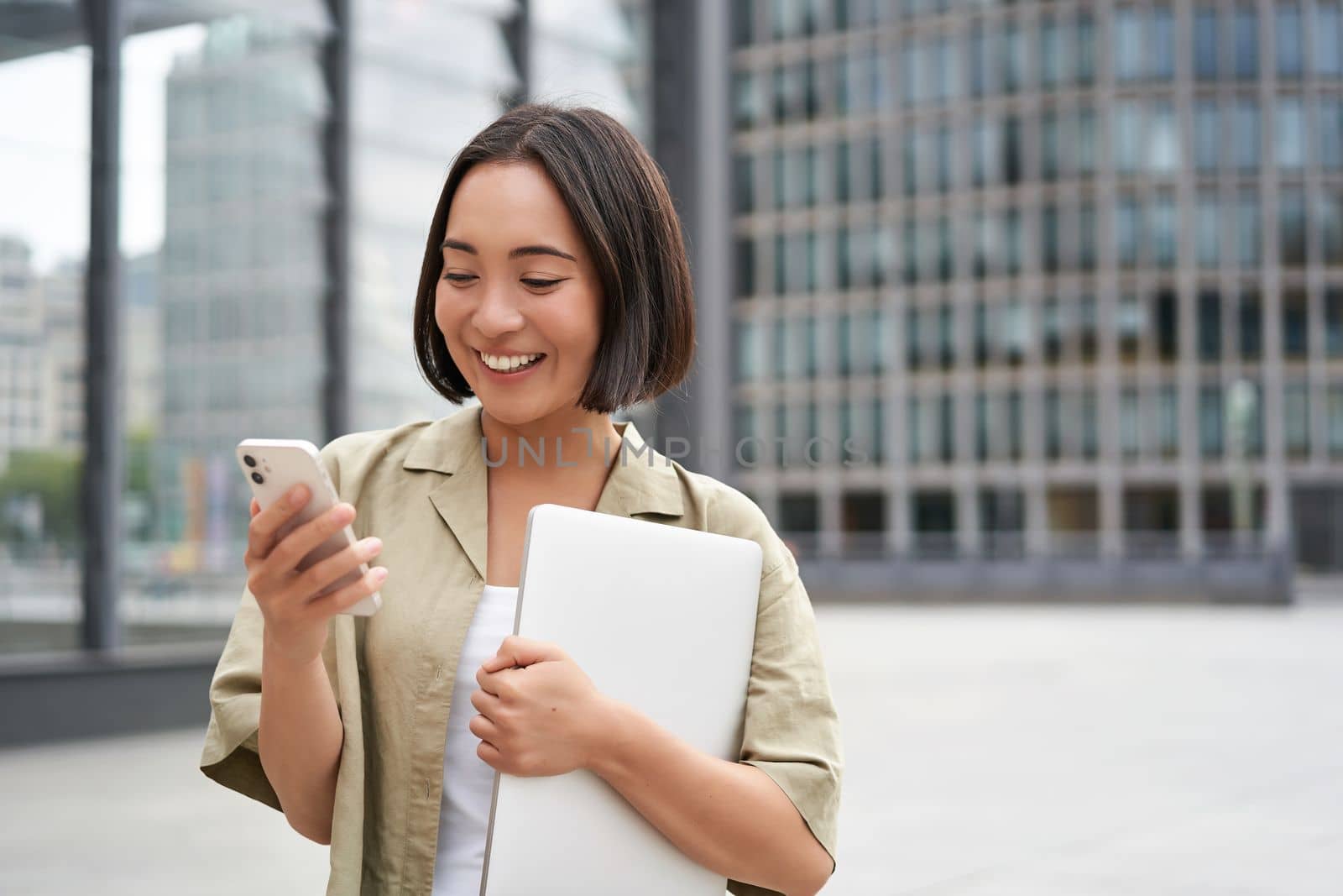 Portrait of young smiling asian woman walking on street, going to work with laptop and smartphone.