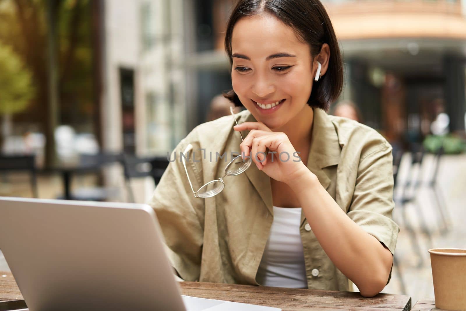 Smiling young korean woman looks at her laptop screen with pleased face, works remotely from outdoors, drinks coffee.