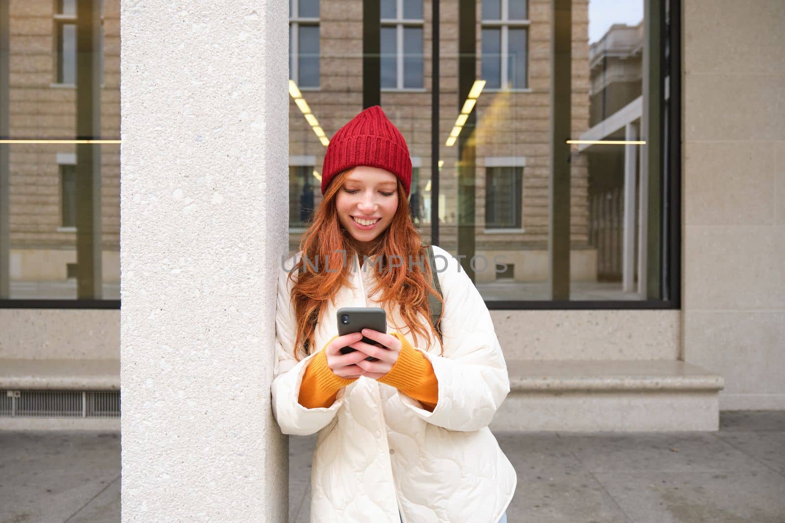 Stylish urban girl using mobile phone app, standing in city, waiting for taxi, looking at smartphone application, texting message.