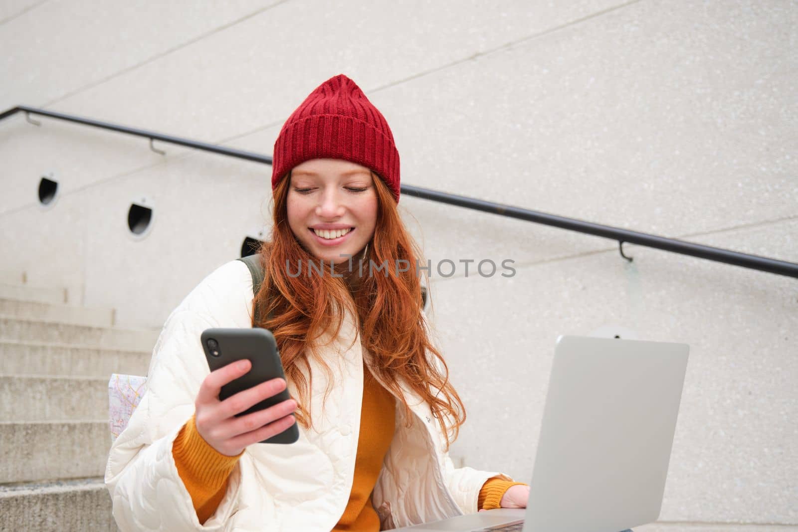 Smiling redhead woman with mobile phone and laptop, sitting on stairs outside building, connects to public wifi, using smartphone and computer.