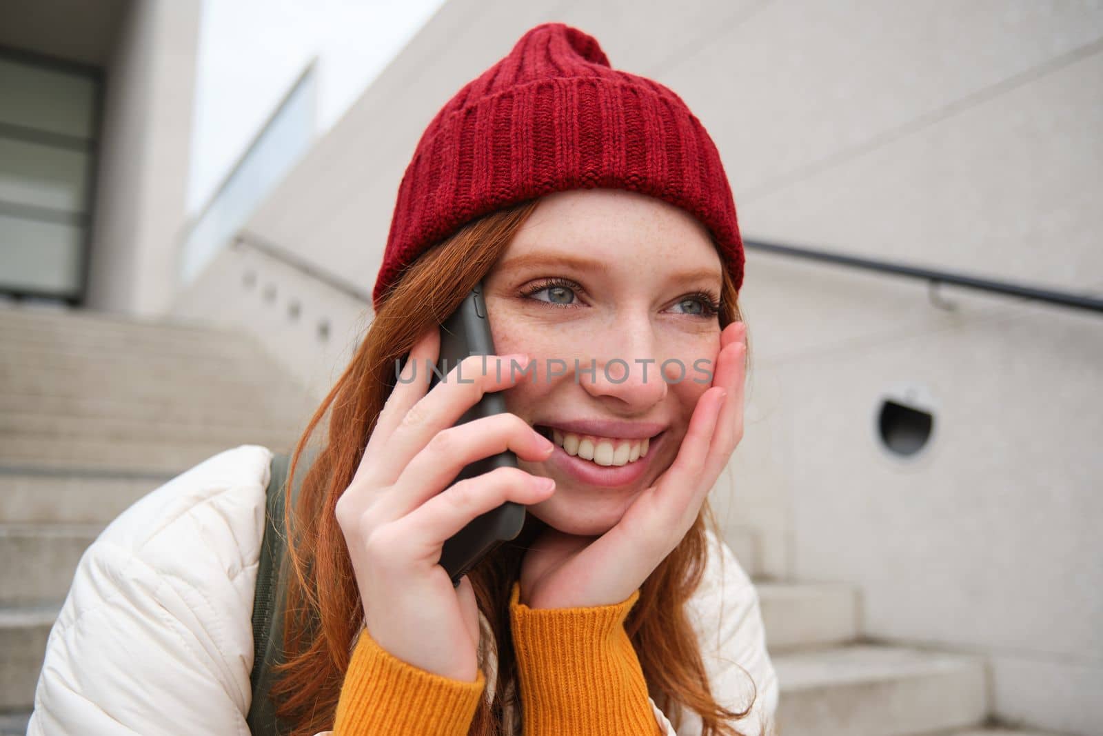 Young stylish redhead girl in red hat, sits on street and talks on mobile phone, has telephone conversation, rings her friend while relaxes outdoors by Benzoix