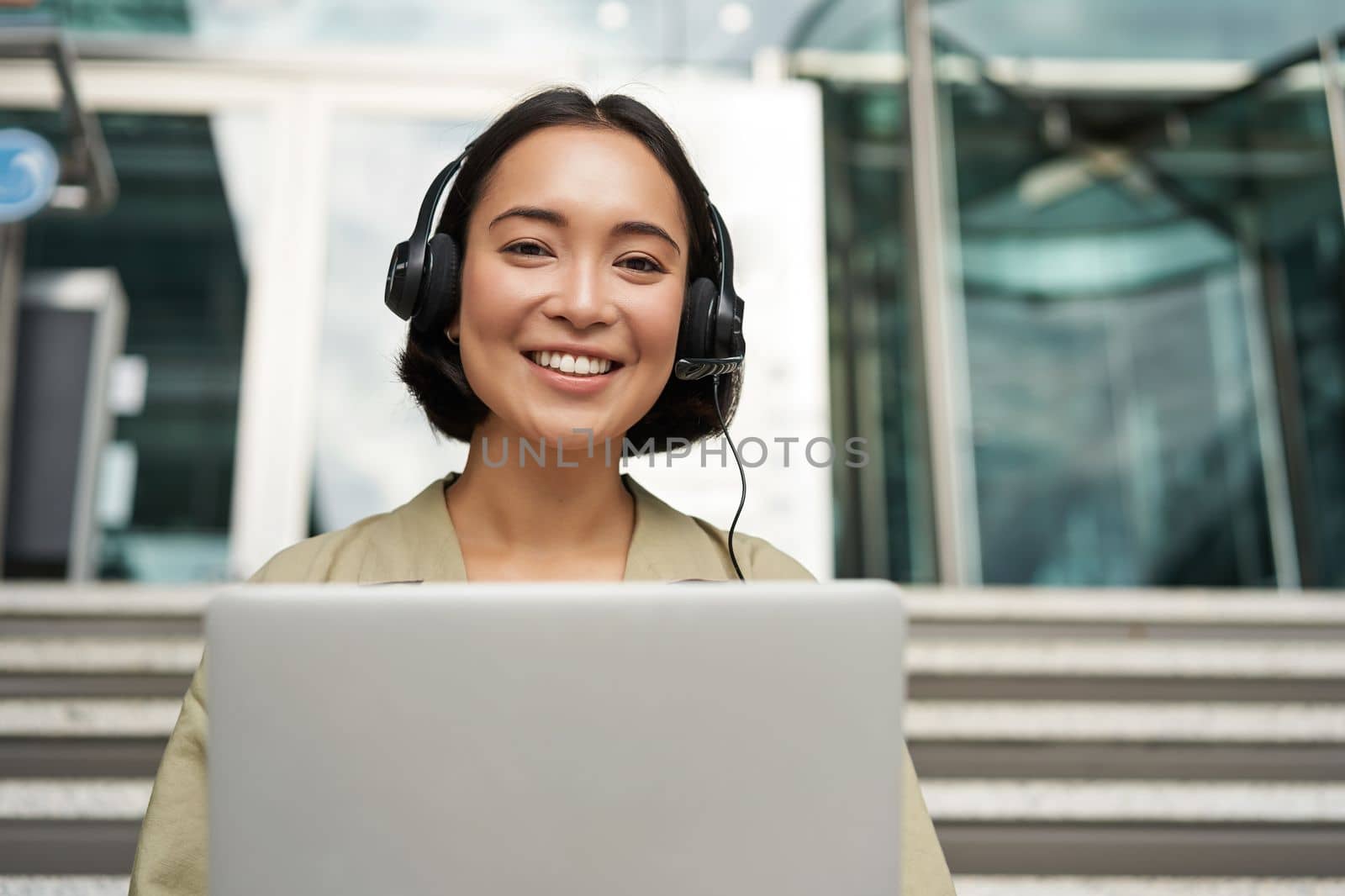 Portrait of young asian woman sitting with laptop and headphones, watching video, does online course on computer, sitting on stairs outdoors.