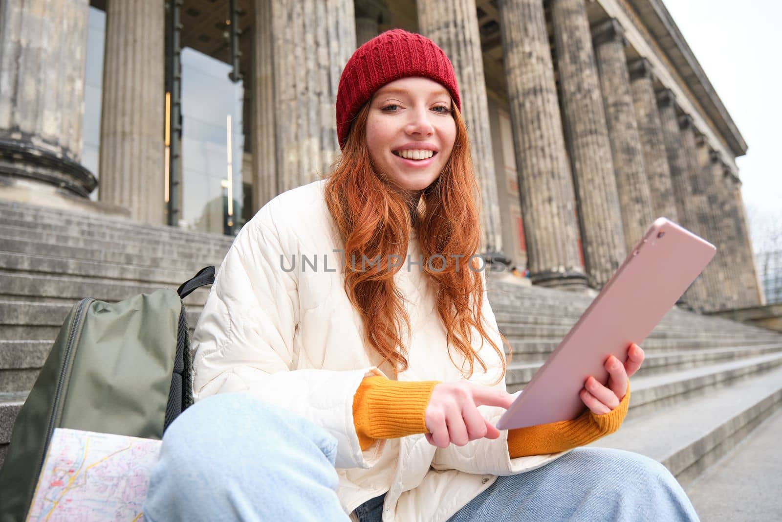 Outdoor shot of young stylish redhead girl sits on staircase and connects to public wifi, uses digital tablet, reads news on gadget by Benzoix