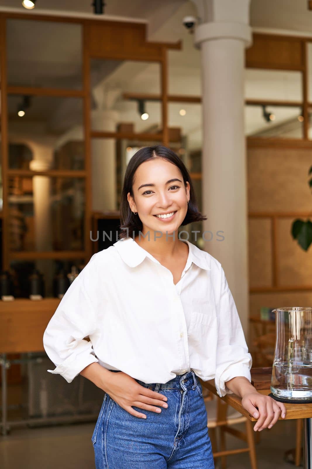 Smiling asian manager, confident woman standing near restaurant entrance, cafe owner welcomes guests.