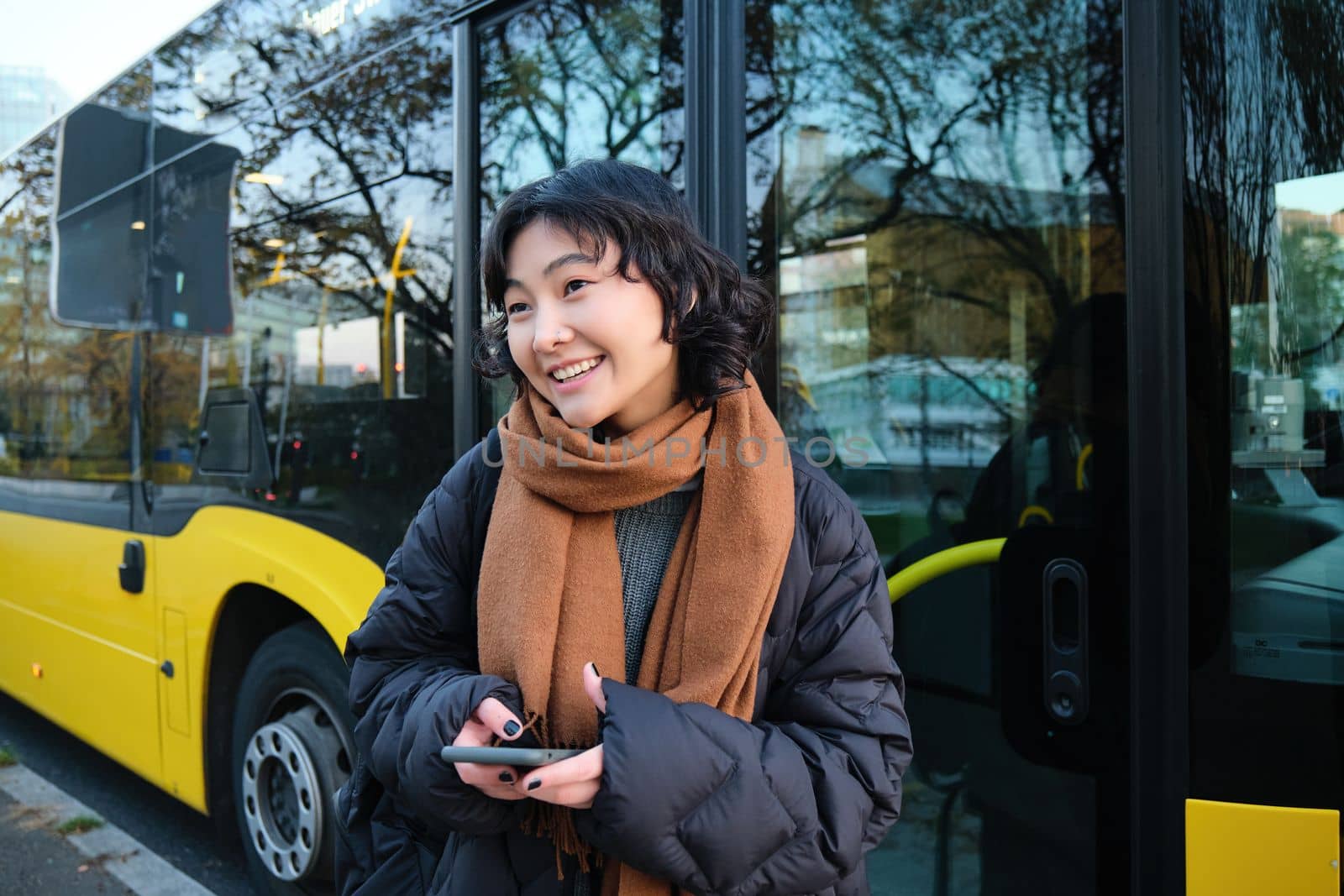 Young beautiful woman standing on bus stop, texting message on smartphone, holding mobile phone, checking her schedule, buying ticket online, wearing winter clothes.