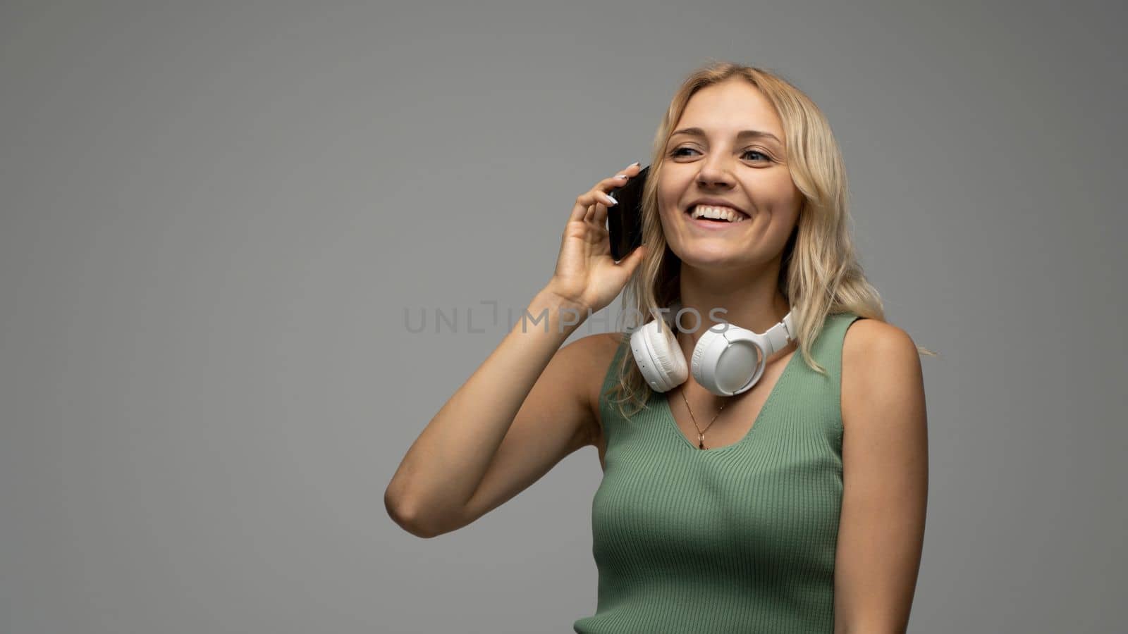Young smiling friendly fun student happy woman 20s in casual green t-shirt with white headphones on a shoulders and speaks on mobile phone