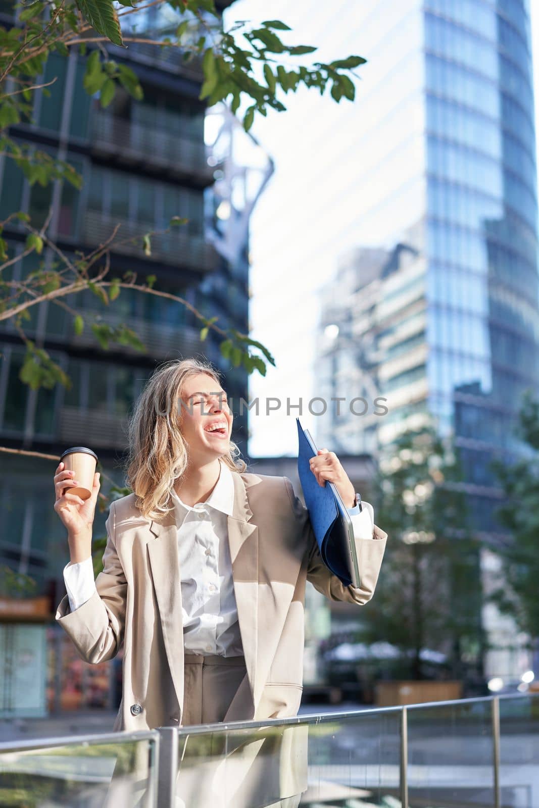 Beautiful businesswoman feels excited, drinks coffee takeaway near business center outside, stands on city street and smiles.
