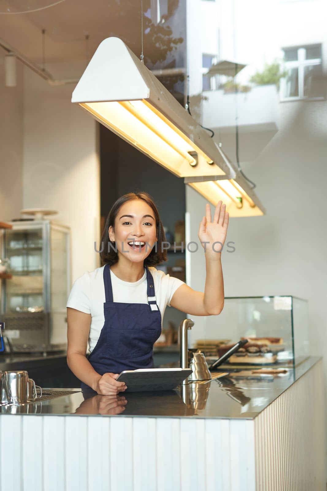 Happy asian girl barista works in coffee shop, waves at client, processing orders behind counter in cafe, wears blue apron.