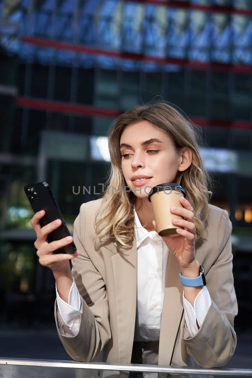 Vertical shot of businesswoman drinks coffee, looks at mobile phone app. Corporate woman on her lunch break, using smartphone.