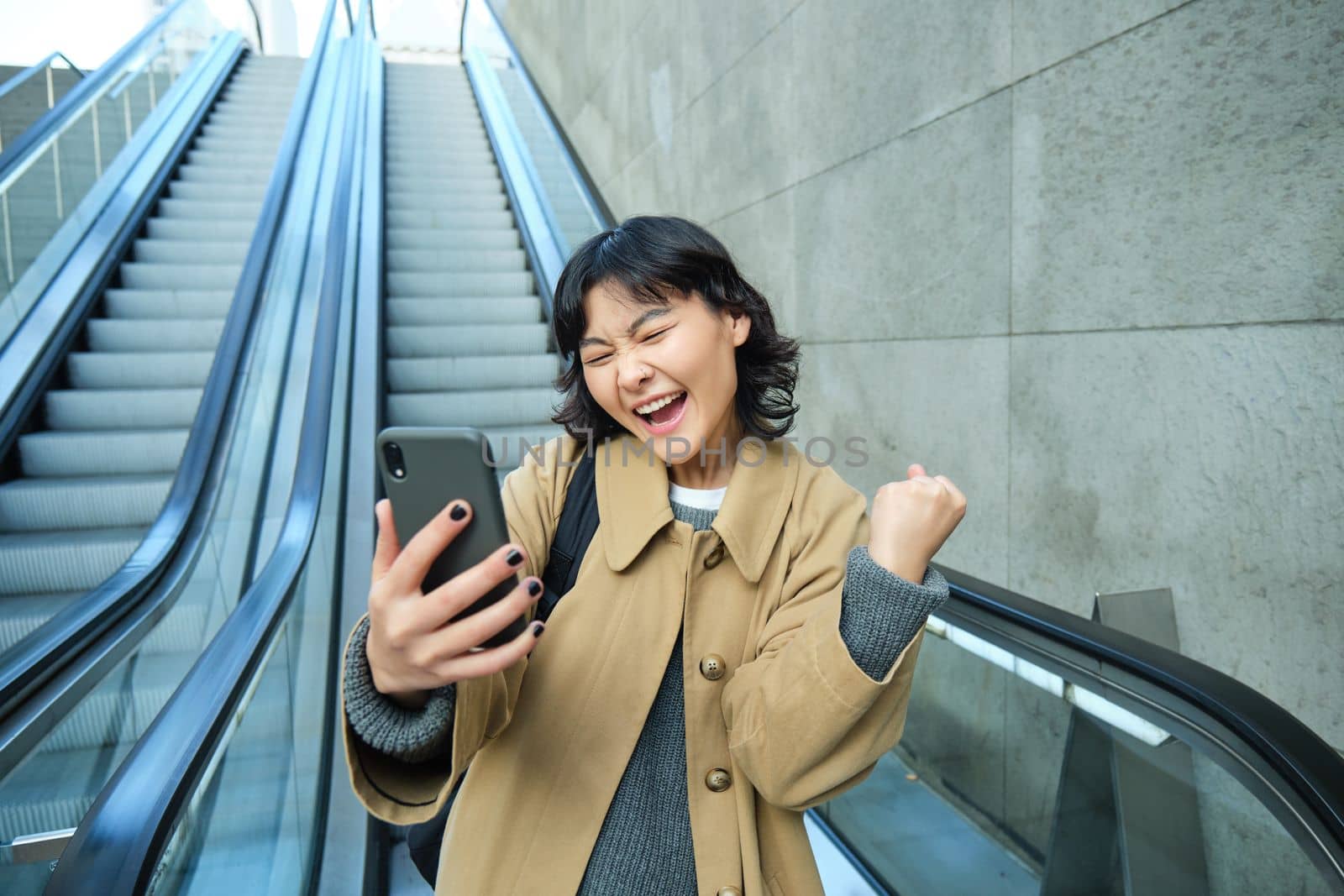 Happy triumphing asian girl, looks amazed and joyful, holds smartphone, goes down escalator with pleased face, wins, hears good news.
