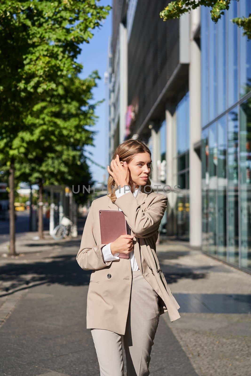 Vertical shot of successful business woman, team leader in suit, holding digital tablet, walking on street. Corporate lady goes to work.