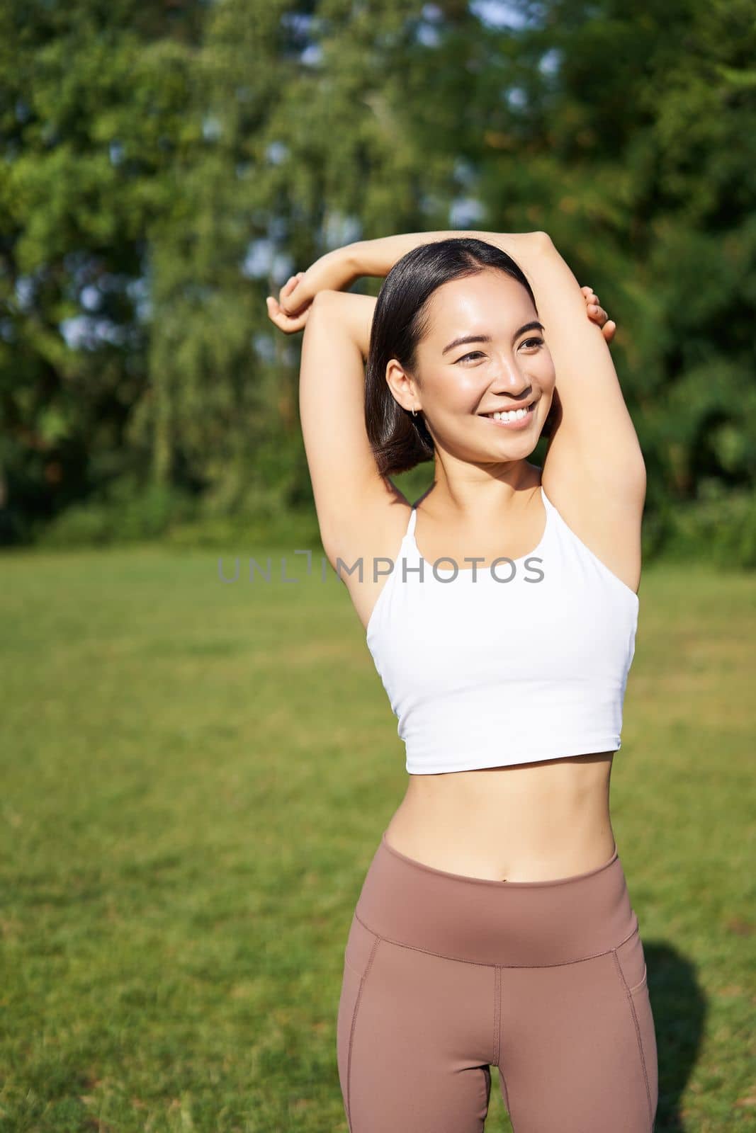 Asian woman stretching her arms, doing fitness workout in park, smiling pleased, warming up before jogging on fresh air in daytime by Benzoix