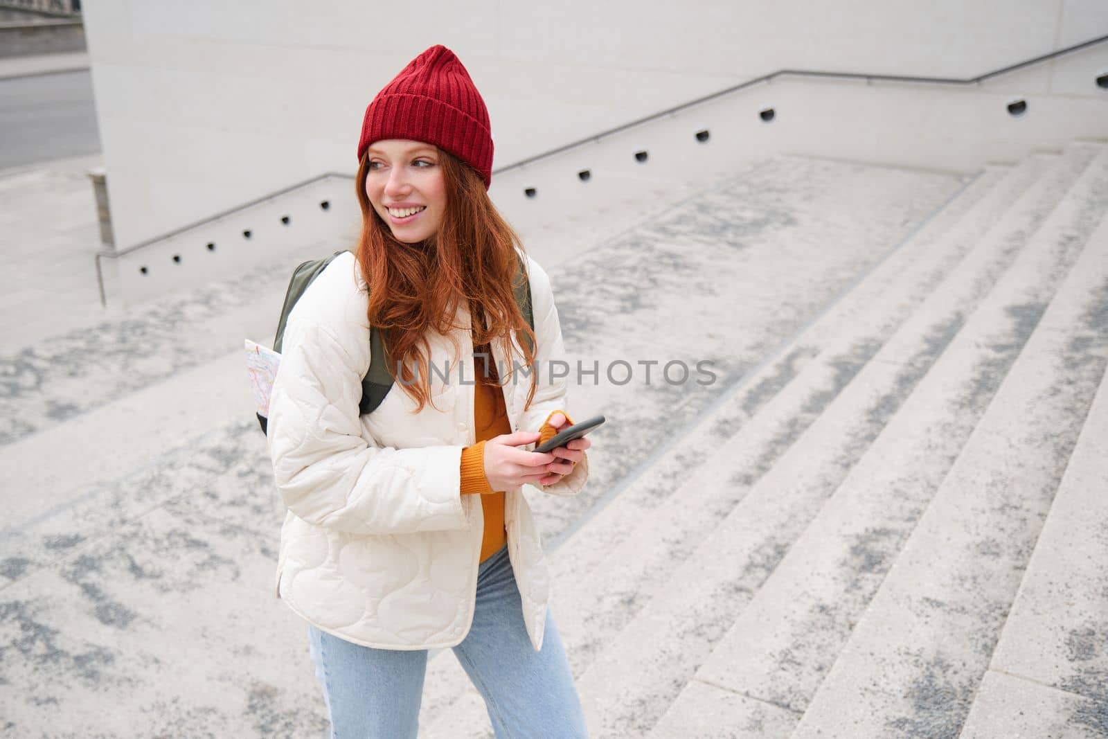 Joyful girl tourist looks at mobile phone, texts message on smartphone social media application, walks around city, looks for sightseeing on mobile app by Benzoix