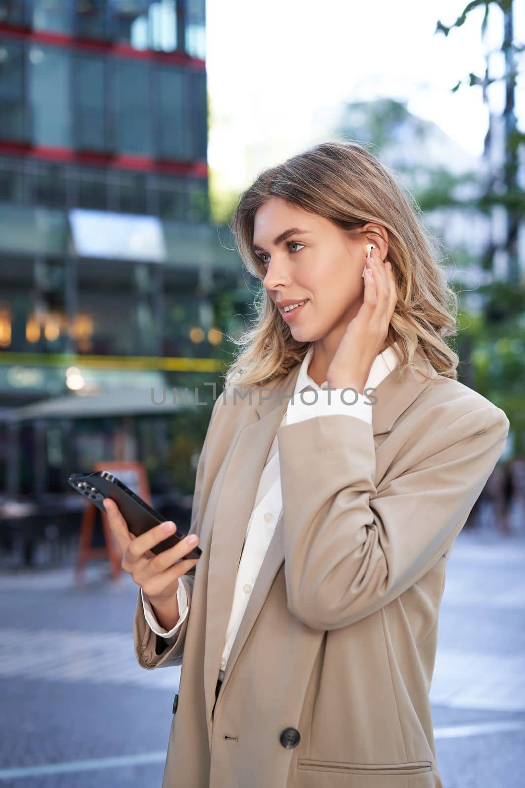 Vertical shot of businesswoman in suit, listening music in wireless headphones, holding smartphone, standing outdoors.