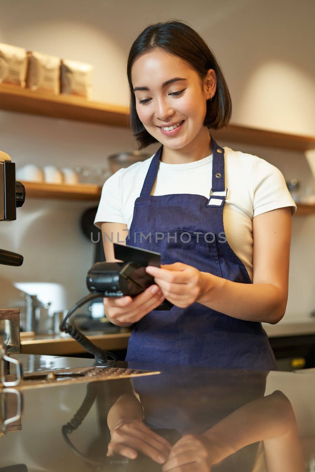 Beautiful smiling asian girl, barista insert client credit card in POS terminal, processing payment, taking orders in coffee shop.