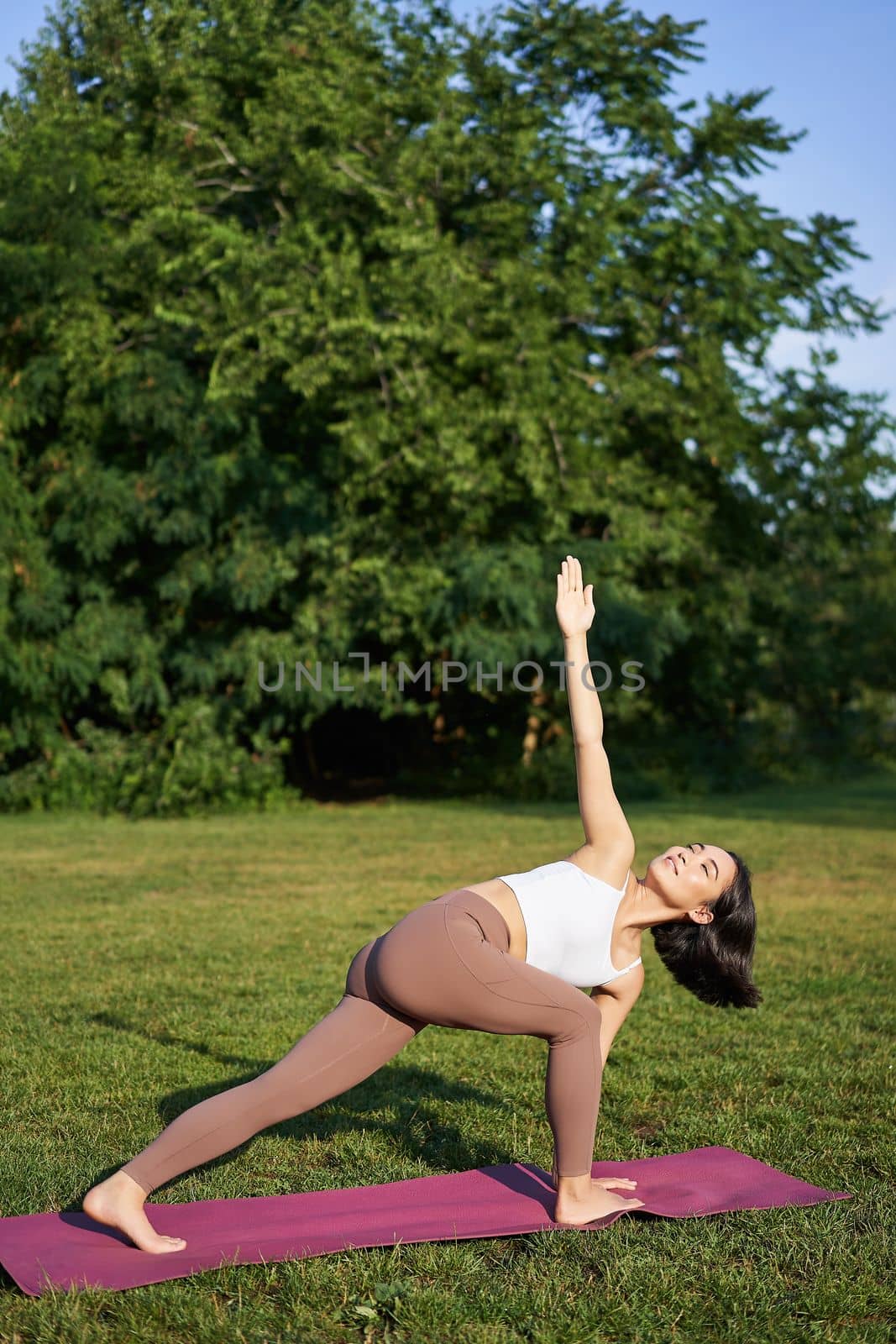 Vertical shot of young korean woman doing yoga training on rubber mat, making asana exercises on fresh air in park by Benzoix