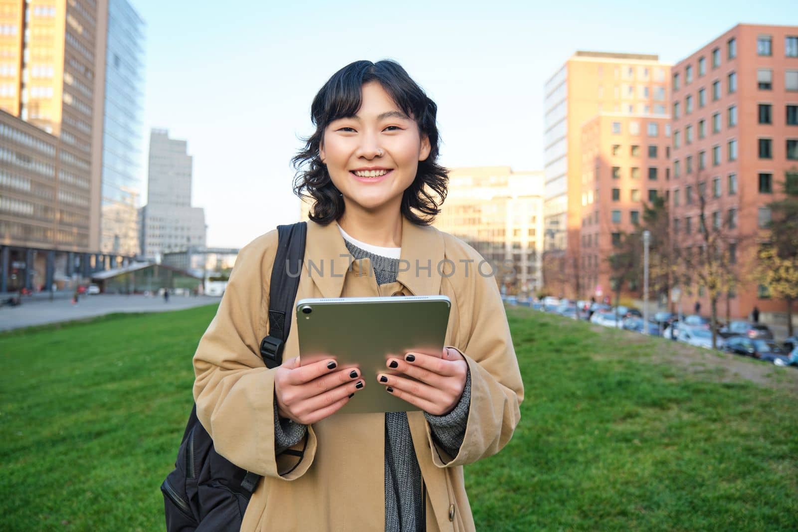 Happy asian girl stands on street, university student walks with digital tablet in hands and smiles, stands in city centre.