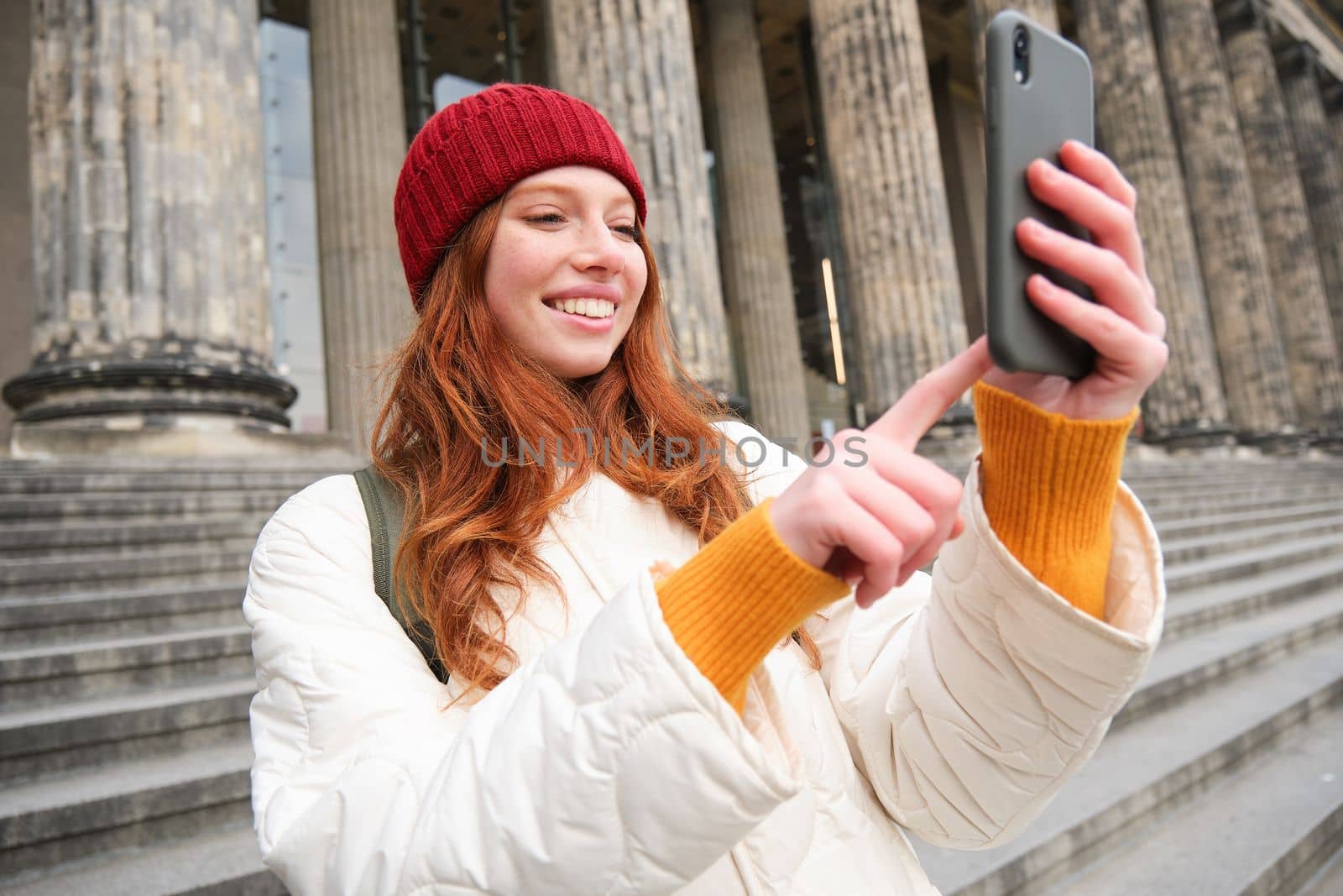 Portrait of young blogger, redhead girl tourist takes pictures of sightseeing, makes photos on smartphone camera, records video and smiles by Benzoix