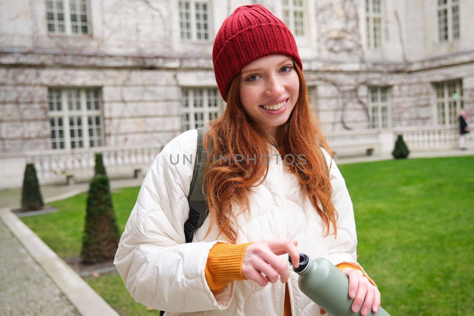 Portrait of happy girl traveler, drinking hot tea from thermos while walking and exploring city. Tourist with flask smiling at camera by Benzoix