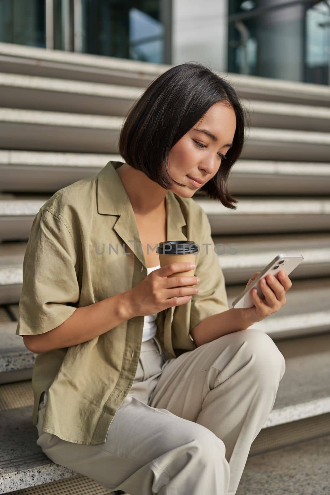 Vertical shot of asian girl drinking her coffee and using mobile phone. Young woman with smartphone and takeaway sits on stairs by Benzoix