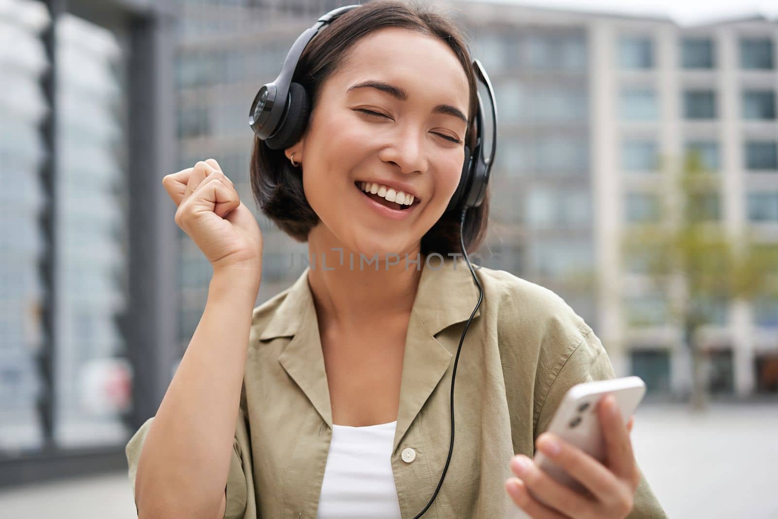 Happy girl dances on street and listens music in wireless headphones, holds mobile phone, using streaming app.