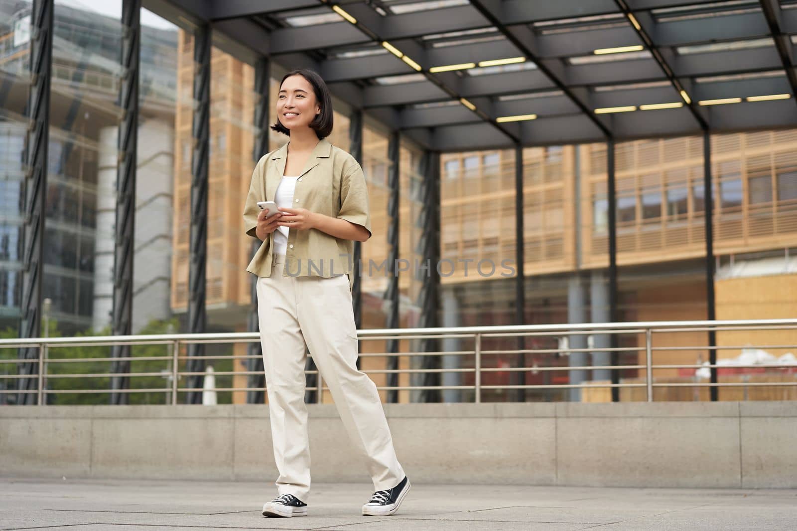 Portrait of young asian woman, student with smartphone, standing on street near glass building, waiting for someone.