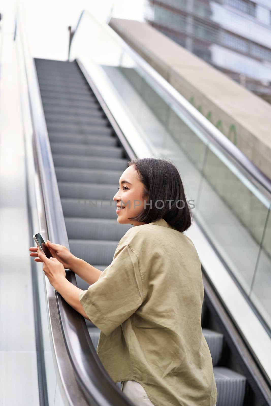 Young happy woman standing on escalator with smartphone, going up, walking in city by Benzoix
