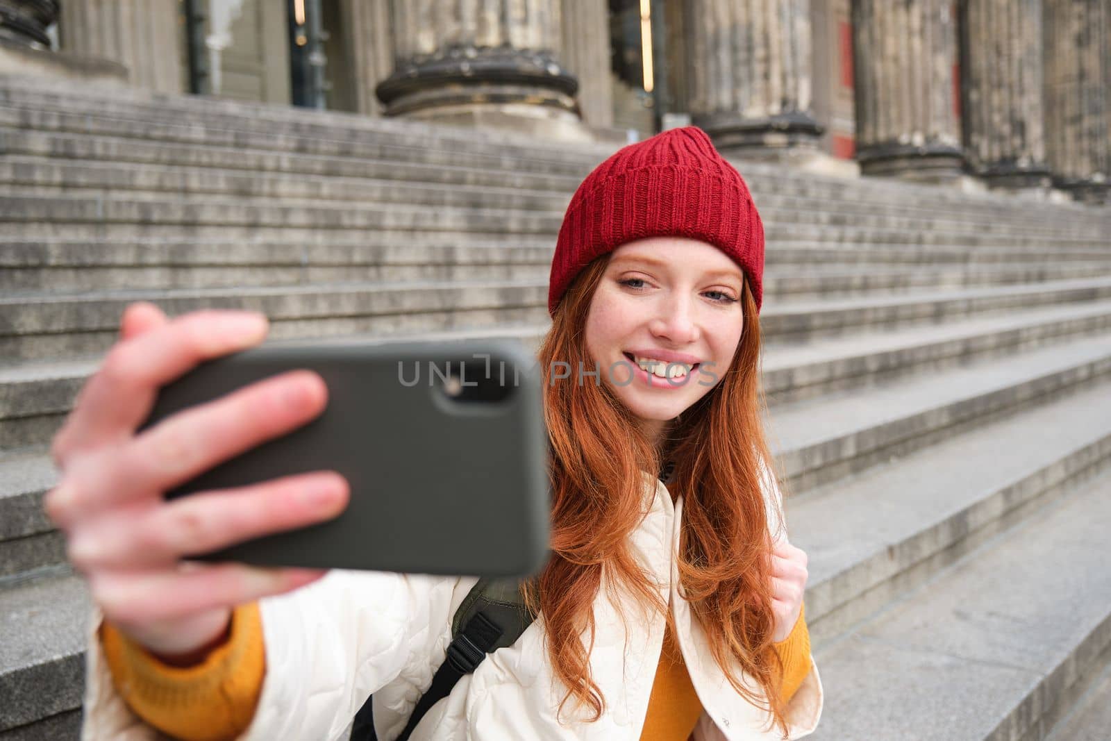 Young redhead tourist takes selfie in front of museum on stairs, holds smartphone and looks at mobile camera, makes photo of herself with phone.