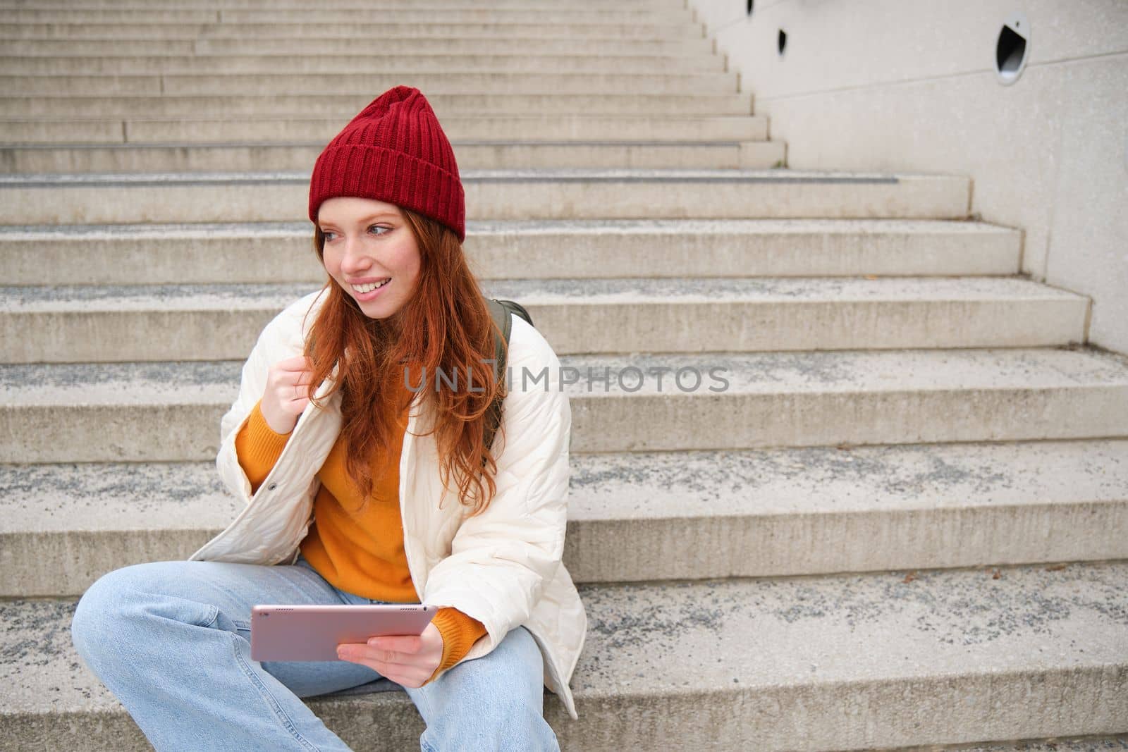 Young beautiful girl with long red hair, smiles, uses internet application, connect to wifi near campus, sits on stairs with digital tablet.