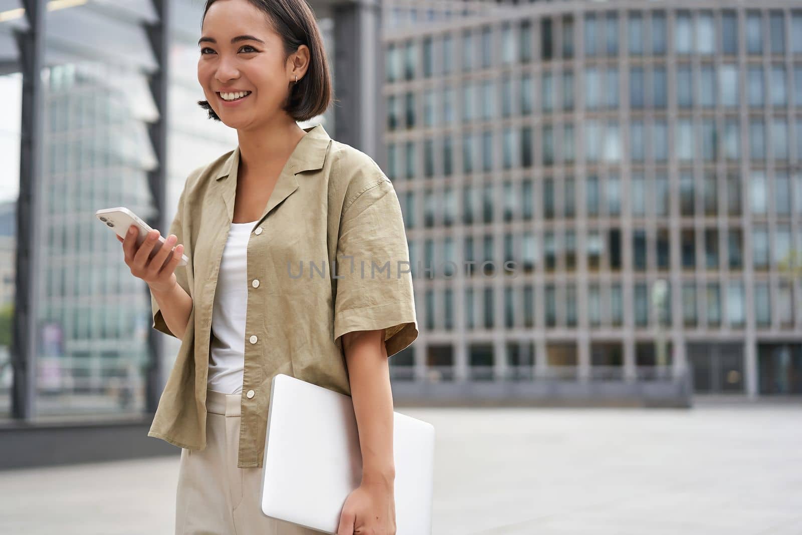 Portrait of asian girl with laptop, going to work or uni, holding smartphone and smiling, walking on street.
