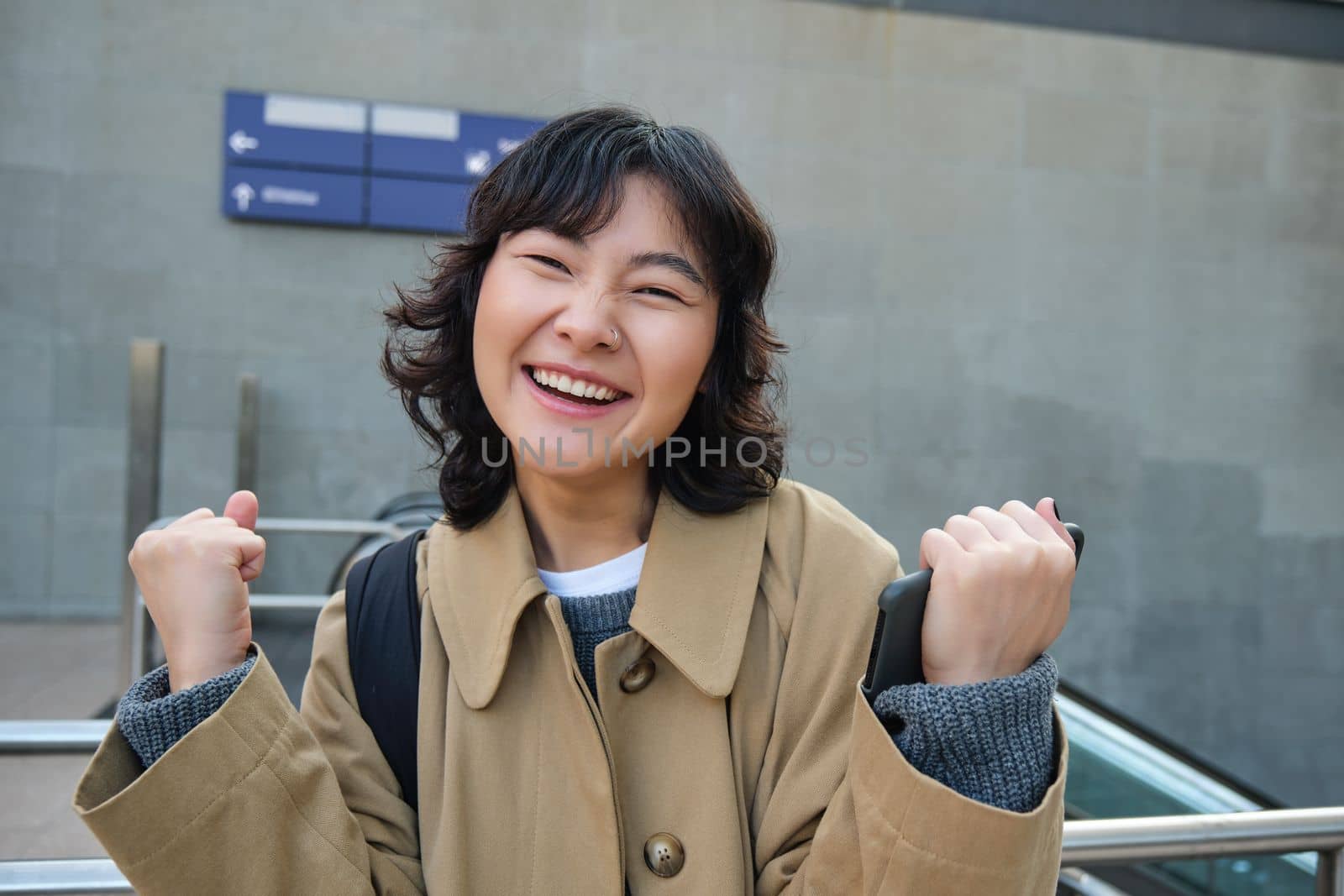 Cheerful asian girl in trench coat, winning, celebrating victory, standing on street and triumphing, smiling happy.
