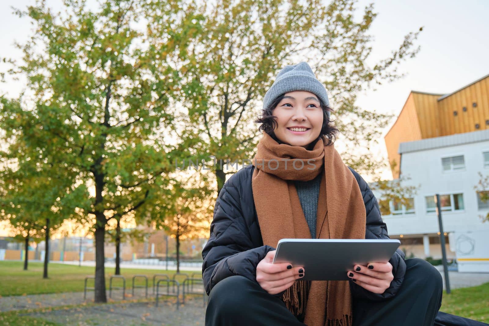 Portrait of asian girl in warm clothes, sits on bench with digital tablet and graphic pen, smiling happily, draws outdoors in chilly weather.