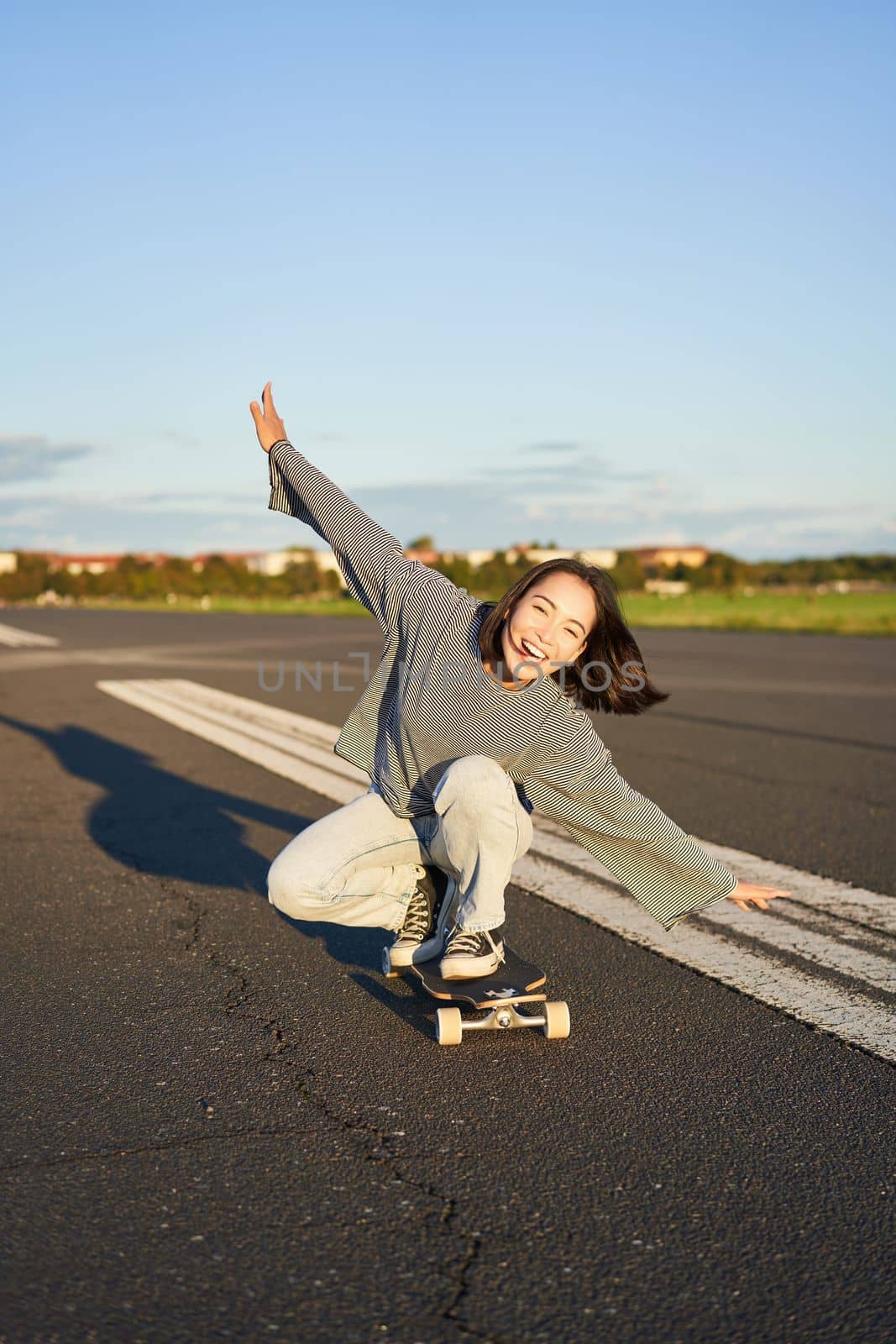 Carefree skater girl on her skateboard, riding longboard on an empty road, holding hands sideways and laughing by Benzoix