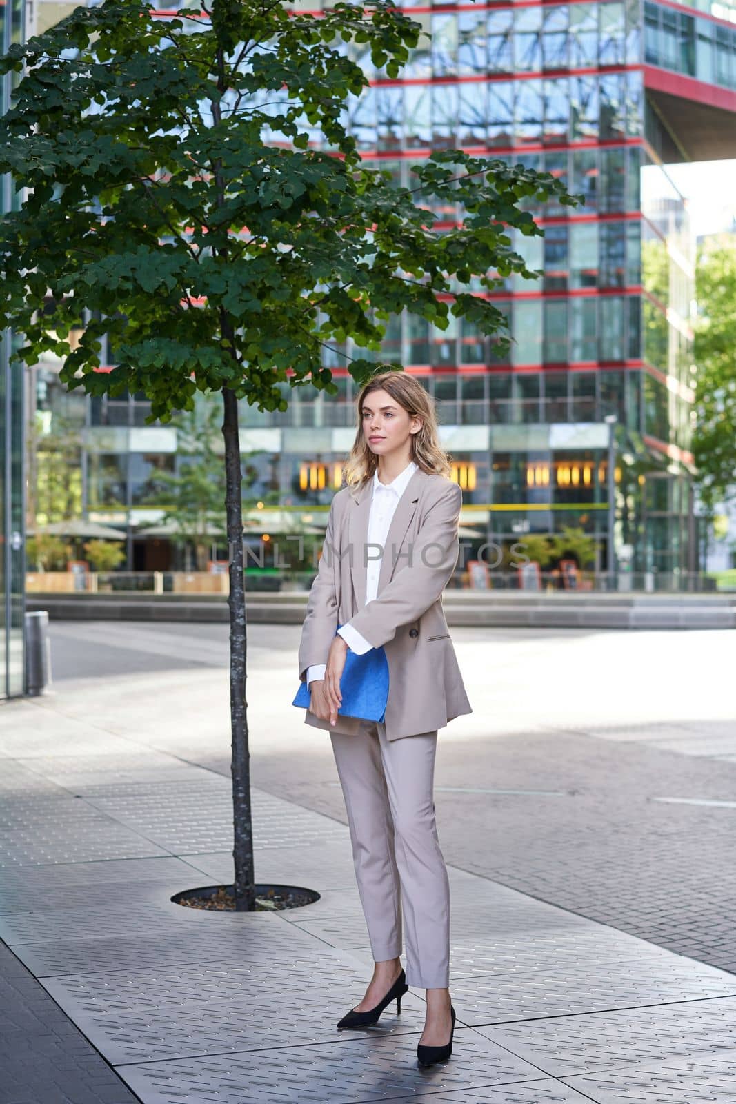 Portrait of young saleswoman in beige business suit, holding blue folder with work documents, standing outdoors on street of city center by Benzoix