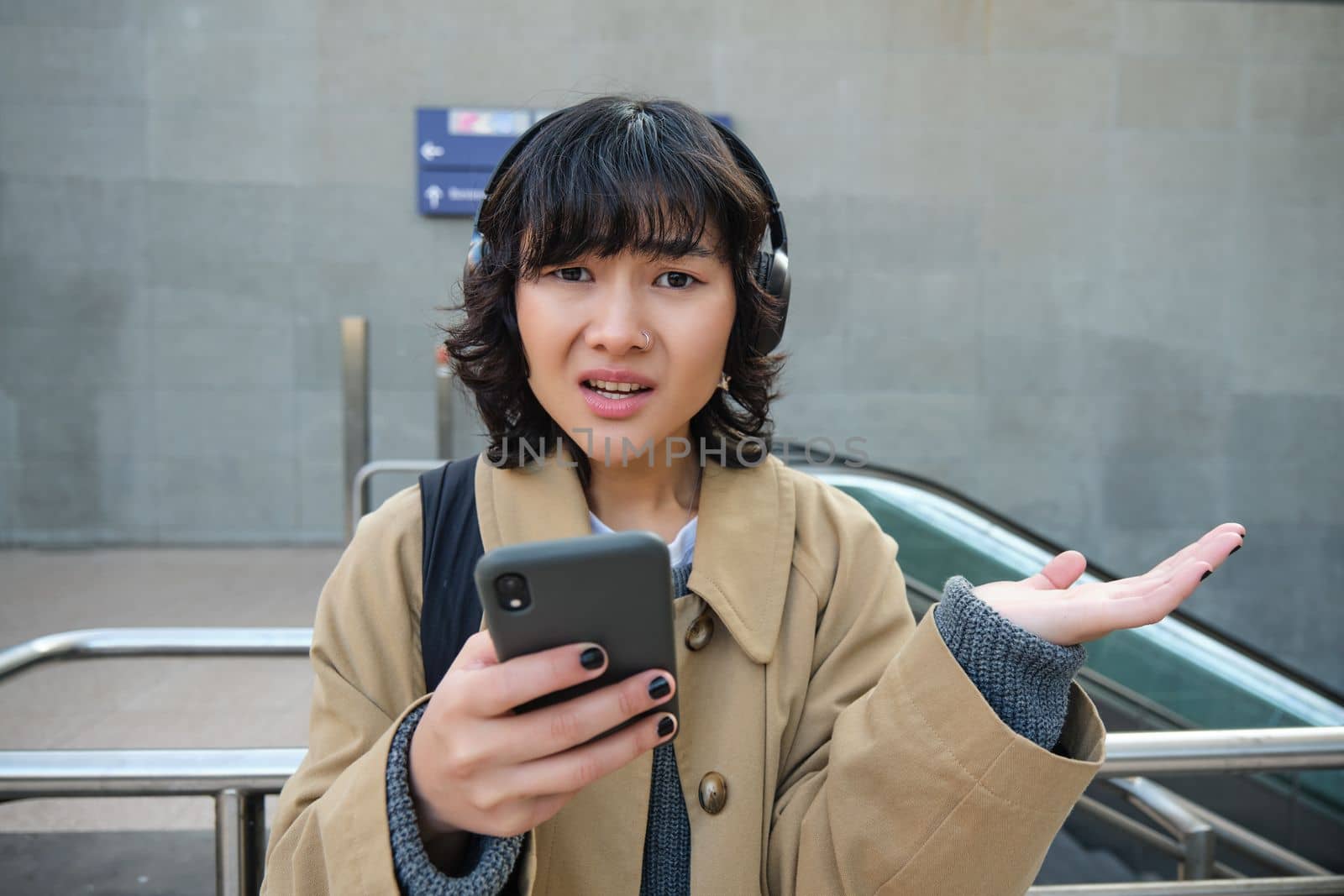 Close up portrait of puzzled, complicated korean girl in headphones, frowns and looks unsure, stands with smartphone and looks at camera while shrugs on street.