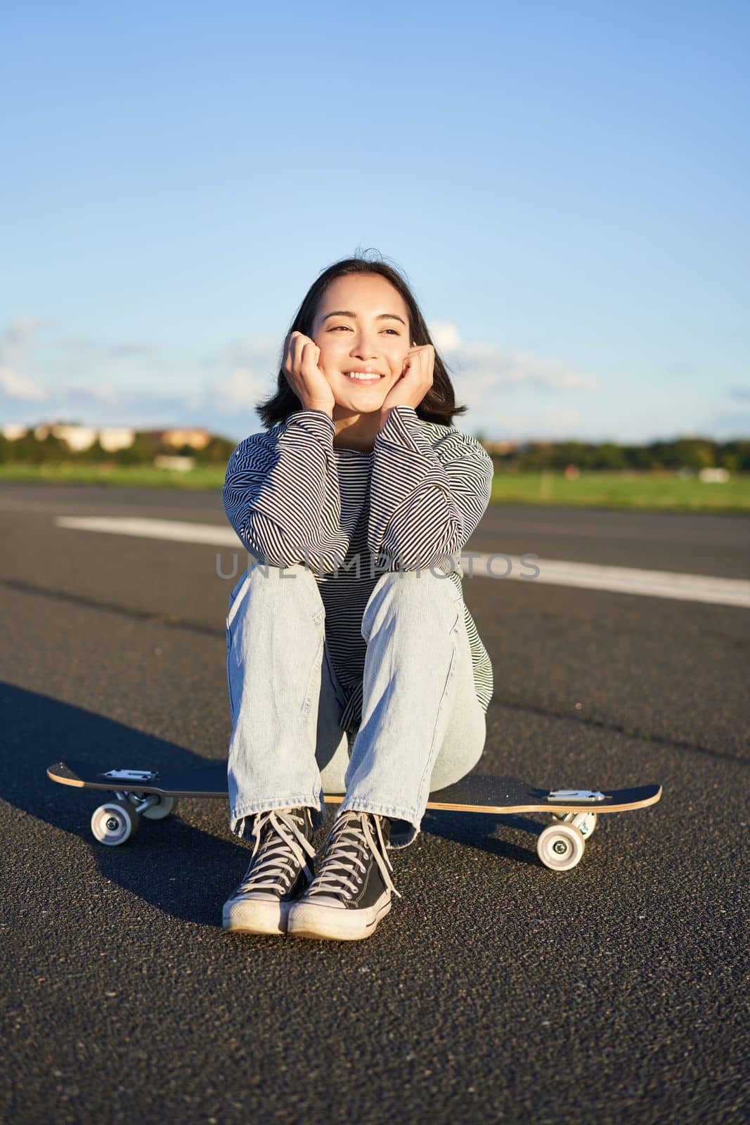Vertical shot of asian girl skater, sits on her skateboard and smiles, enjoys sunny day, cruising on longboard on empty road outdoors by Benzoix