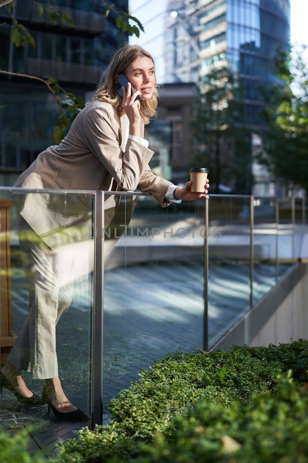 Stylish office woman in beige suit, talking on mobile phone while standing outside on street with morning coffee.