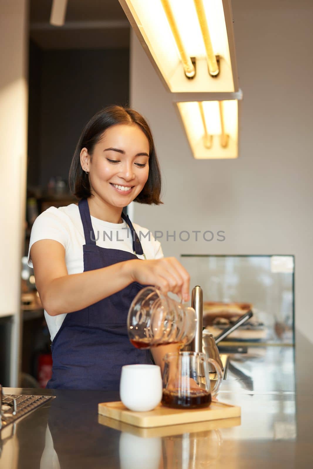 Vertical shot of smiling girl working as barista, prepare pour over, making filter coffee batch brew, standing at counter in cafe in apron.
