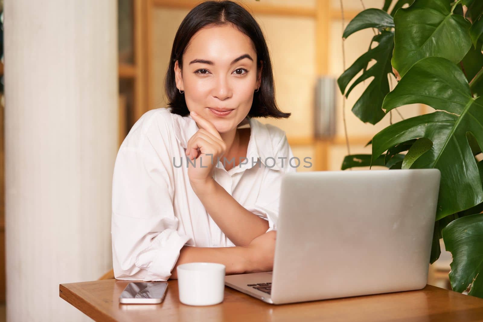Confident young stylish woman with laptop, sitting in cafe and working, freelancer in co-working space with cup of coffee by Benzoix